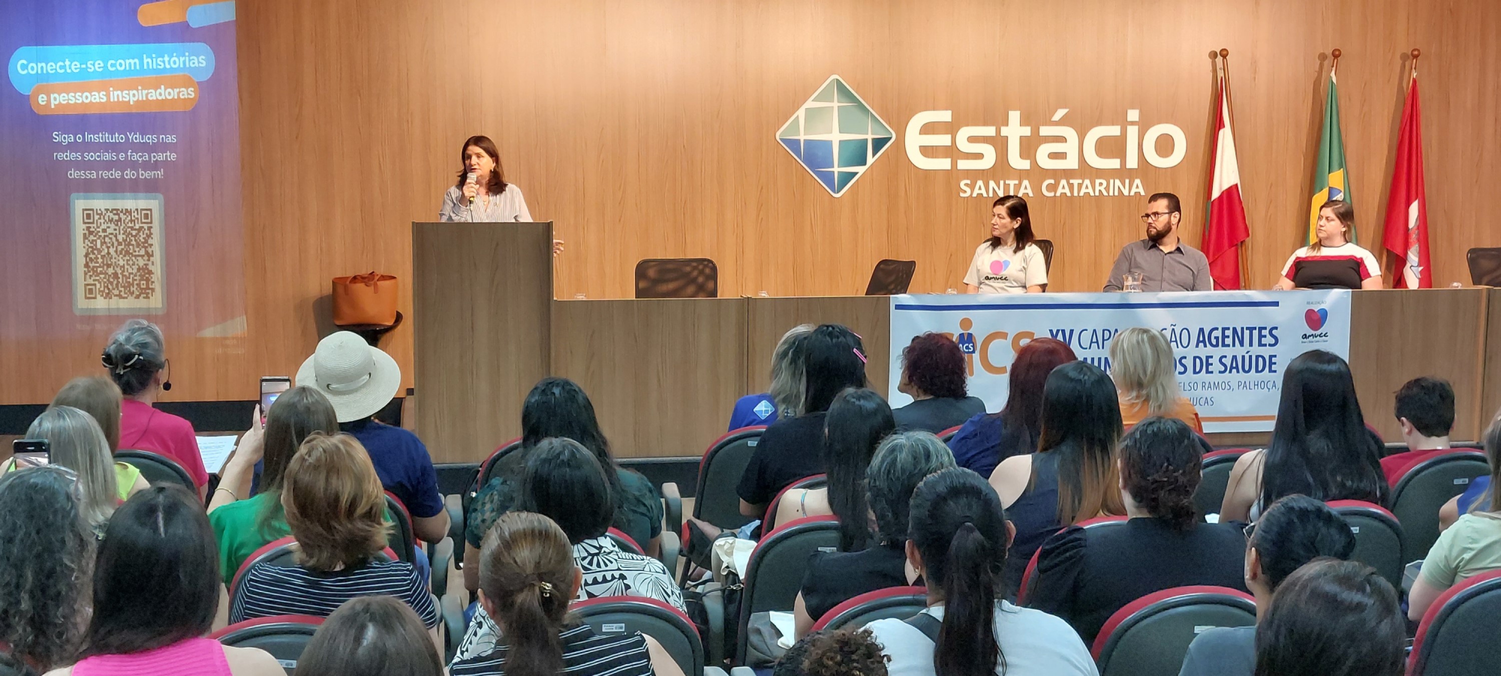 A imagem mostra um auditório durante um evento na Estácio Santa Catarina. No palco, há quatro pessoas sentadas atrás de uma mesa — uma mulher em pé fala ao microfone enquanto três pessoas (duas mulheres e um homem) estão sentadas, ouvindo. Atrás deles, na parede de madeira, está o logotipo da instituição e bandeiras posicionadas à direita. Na lateral esquerda, um telão exibe um slide com um QR code e a frase “Conecte-se com histórias e pessoas inspiradoras”, incentivando a seguir o Instituto Yduqs nas redes sociais. O público está sentado em cadeiras de encosto cinza e borda vermelha, de frente para o palco. Um banner à frente da mesa indica que se trata da “XV Capacitação Agentes Comunitários de Saúde” realizada pela AMUCC.