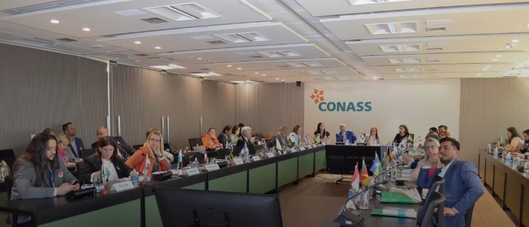 Participantes sentados em mesa em formato de U durante a 10ª reunião do CONASS, com bandeiras dos estados posicionadas à frente.