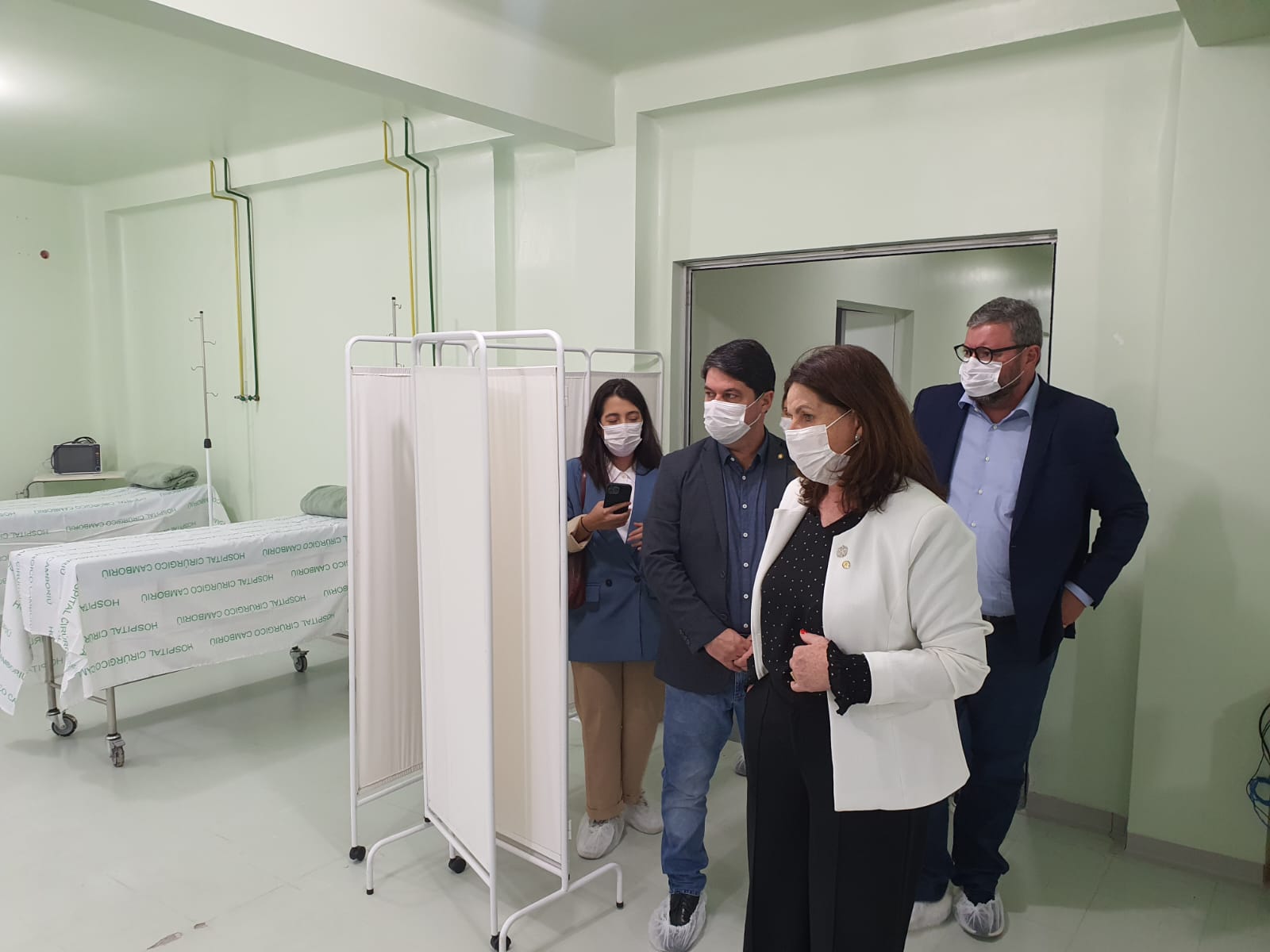 Visita técnica a um hospital, com a secretária de saúde Carmen Zanotto e outros gestores públicos utilizando máscaras e protetores de calçado. Ao fundo, leitos hospitalares com lençóis estampados com 'Hospital Cirúrgico Camboriú'. A cena ocorre em uma enfermaria, com divisórias móveis e estrutura clínica visivelmente recém-reformada.
