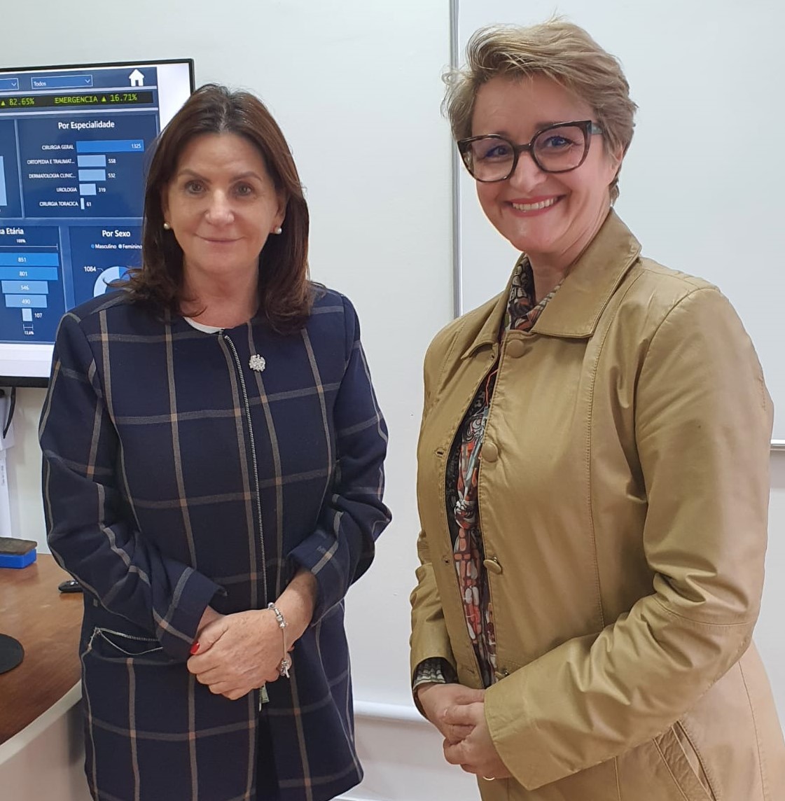 Duas mulheres posam sorrindo para foto em ambiente hospitalar. Ao fundo, tela com gráficos e dados estatísticos relacionados à área da saúde. À esquerda, uma das mulheres veste casaco xadrez azul-marinho; à direita, a outra veste casaco bege claro.