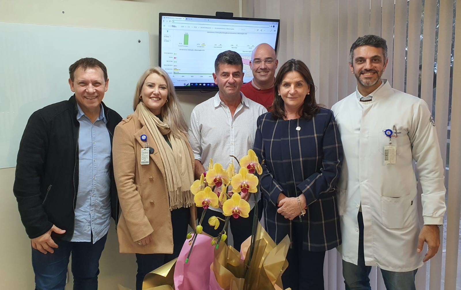 alt='Grupo de seis pessoas posando para foto sorridentes em ambiente hospitalar. Ao fundo, uma televisão exibe gráficos de dados. À frente, um arranjo de orquídeas amarelas com detalhes em rosa, embalado em papel dourado e rosa. Duas mulheres e quatro homens compõem o grupo, incluindo profissionais da saúde e gestores.