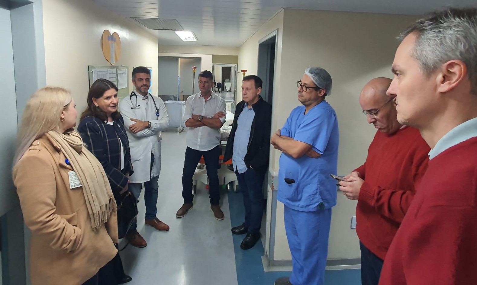 Grupo de profissionais da saúde e representantes reunidos em corredor hospitalar durante visita técnica ao Instituto de Cardiologia. Participantes estão em conversa, alguns de jaleco branco e roupa cirúrgica, demonstrando ambiente clínico em funcionamento.