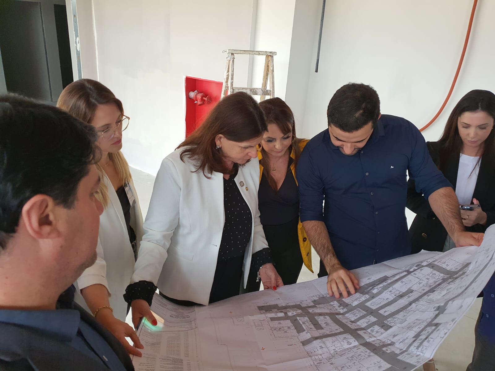 Secretária de Saúde Carmen Zanotto e equipe técnica analisam a planta arquitetônica das obras do Hospital de Itapema. O grupo está reunido dentro da construção, observando detalhes do projeto impresso. O ambiente tem aparência de obra em andamento, com paredes brancas inacabadas, escada de alumínio e tubulações expostas.
