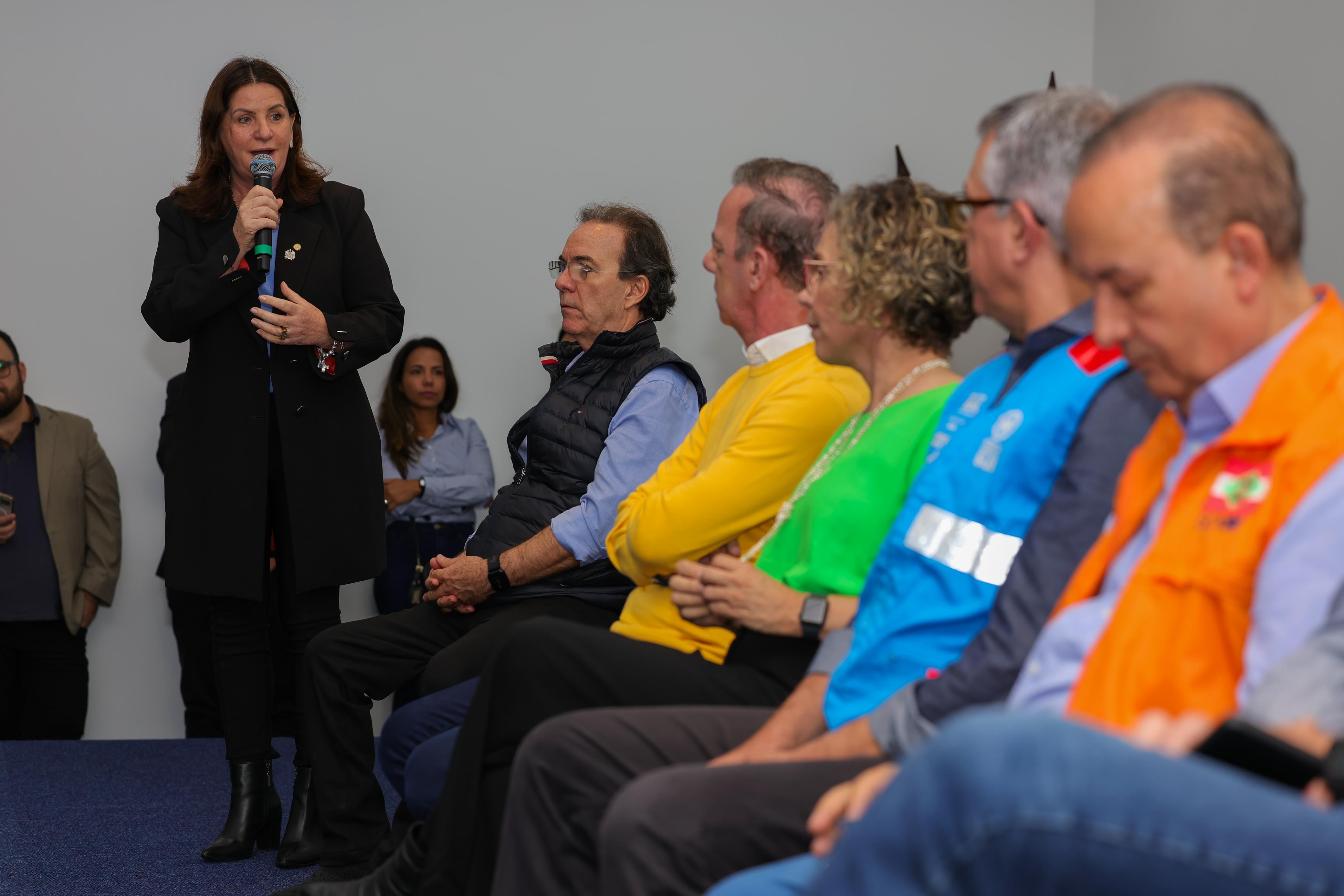 Mulher de pé falando ao microfone para grupo de autoridades sentadas, incluindo representantes do governo e defesa civil, durante reunião no Vale do Itajaí.