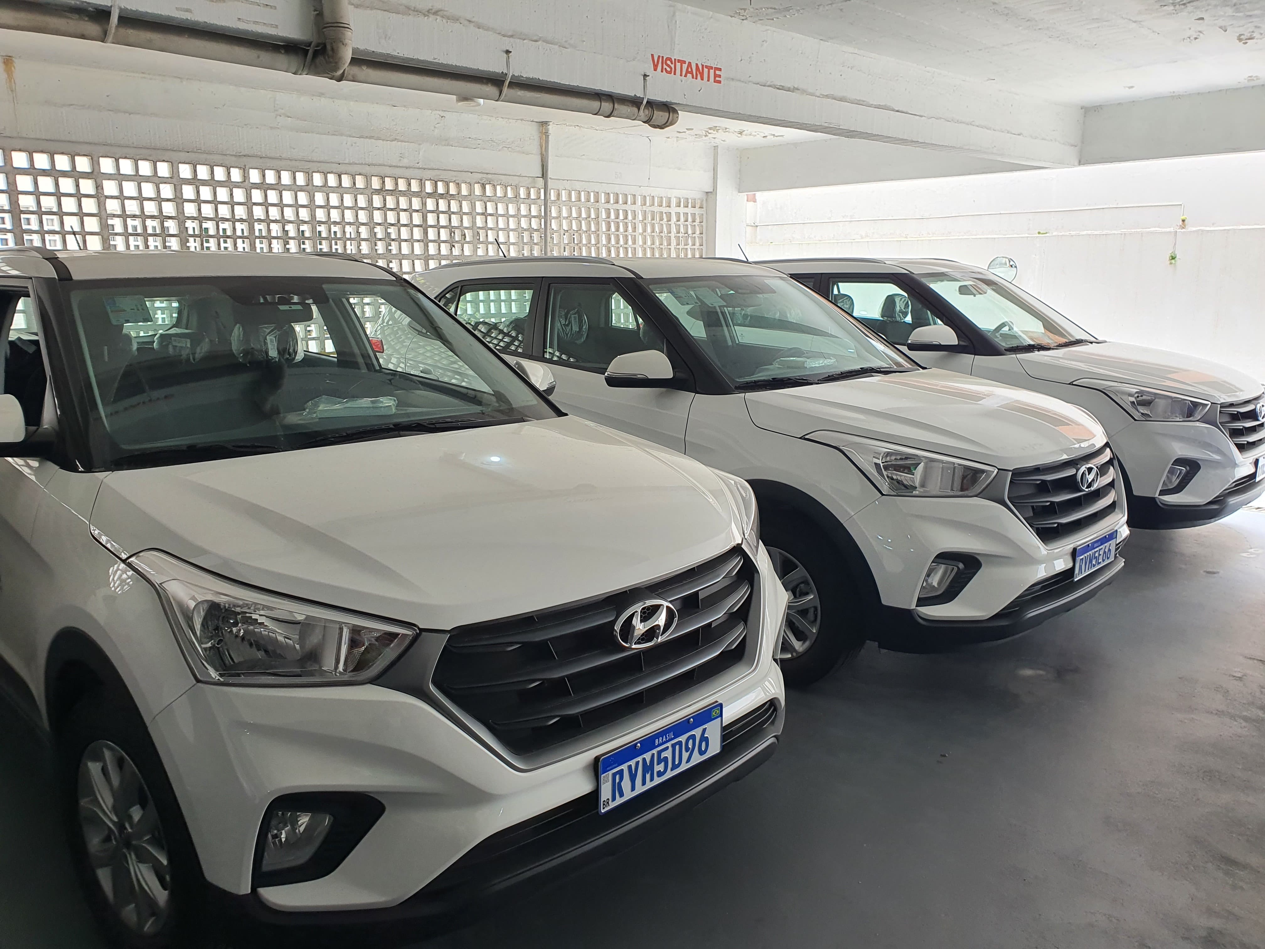 A imagem mostra três carros Hyundai Creta brancos estacionados lado a lado em um estacionamento coberto. As placas são azuis, padrão de veículos oficiais no Brasil, e todos parecem novos, ainda com plásticos de proteção no interior. Na parede, acima dos veículos, há uma pintura em vermelho com a palavra “VISITANTE”. A iluminação natural entra pelas aberturas de tijolos vazados no fundo e pela lateral direita do estacionamento.