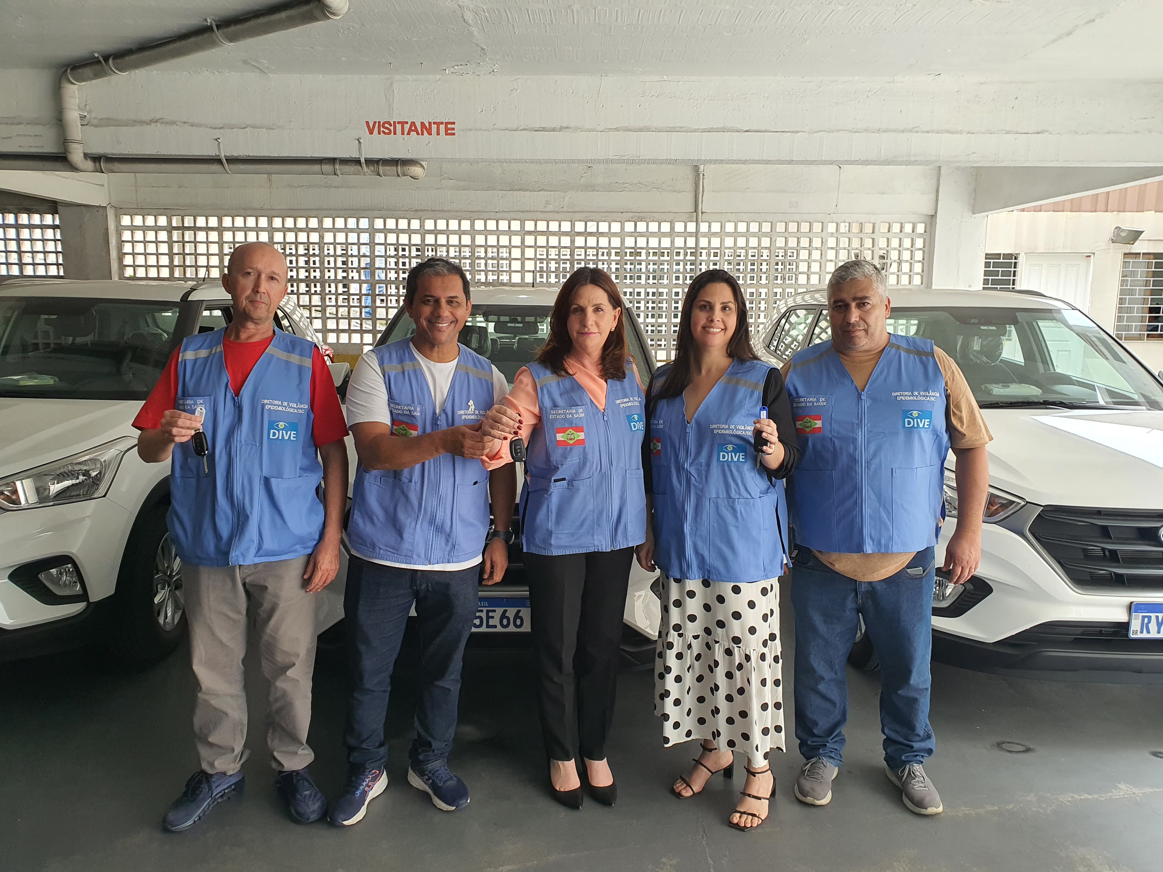 A imagem mostra cinco pessoas posando em frente a três carros Hyundai Creta brancos estacionados em um local coberto. Todos vestem coletes azuis com logotipos do Governo de Santa Catarina, Secretaria de Estado da Saúde e DIVE (Diretoria de Vigilância Epidemiológica). Quatro deles seguram chaves de veículos, sugerindo a entrega oficial dos carros para uso institucional. Na parede atrás, há uma marcação em vermelho com a palavra “VISITANTE”, e o fundo tem elementos vazados que permitem entrada de luz natural.