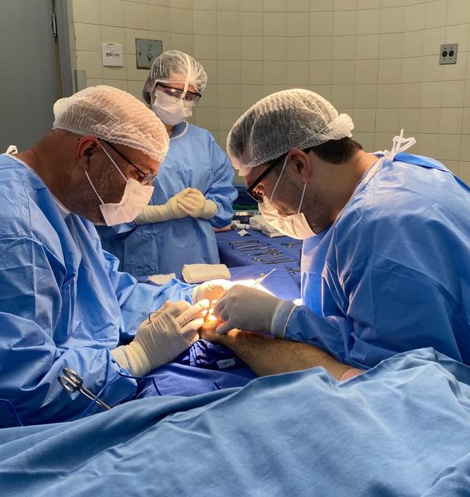 Equipe médica realiza procedimento cirúrgico em um centro cirúrgico hospitalar; dois profissionais concentram-se na operação da mão de um paciente, enquanto uma terceira pessoa observa ao fundo. Todos usam roupas e equipamentos de proteção esterilizados, incluindo toucas, máscaras e luvas.