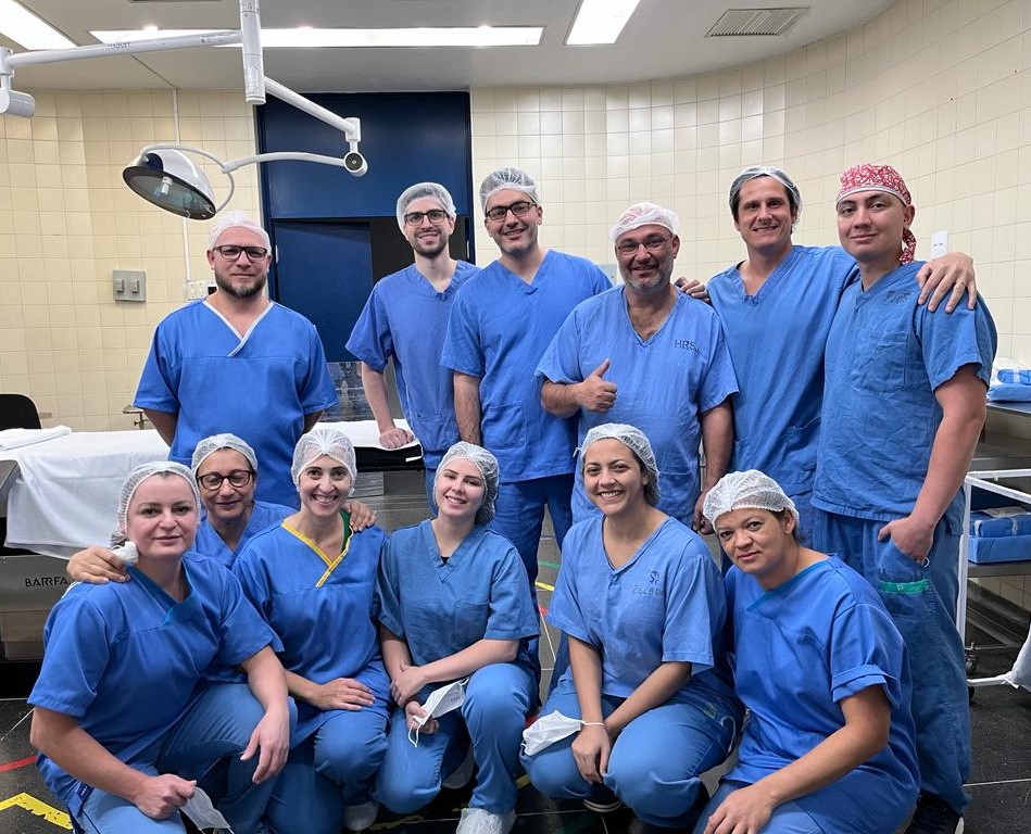 Equipe médica formada por homens e mulheres posa para foto em uma sala cirúrgica. Todos estão usando roupas hospitalares azuis, toucas e alguns com máscaras abaixadas. O ambiente é iluminado, com paredes revestidas de azulejos claros e equipamentos médicos ao fundo. A equipe sorri, demonstrando espírito de colaboração e missão cumprida.
