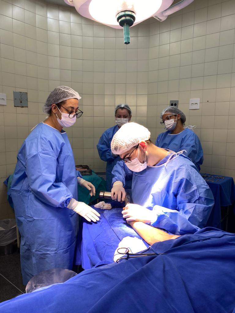 Profissionais da saúde realizam procedimento cirúrgico em uma sala de operação. Um cirurgião trabalha na mão de um paciente deitado, enquanto outros três membros da equipe observam e auxiliam. Todos usam roupas hospitalares esterilizadas, toucas, máscaras e luvas. A iluminação cirúrgica está acesa sobre o campo operatório.
