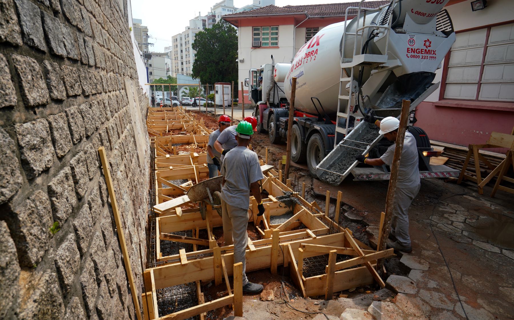 Trabalhadores da construção civil com capacetes despejando concreto em formas de madeira com auxílio de caminhão betoneira da empresa Engemix, próximo a um prédio de fachada vermelha.