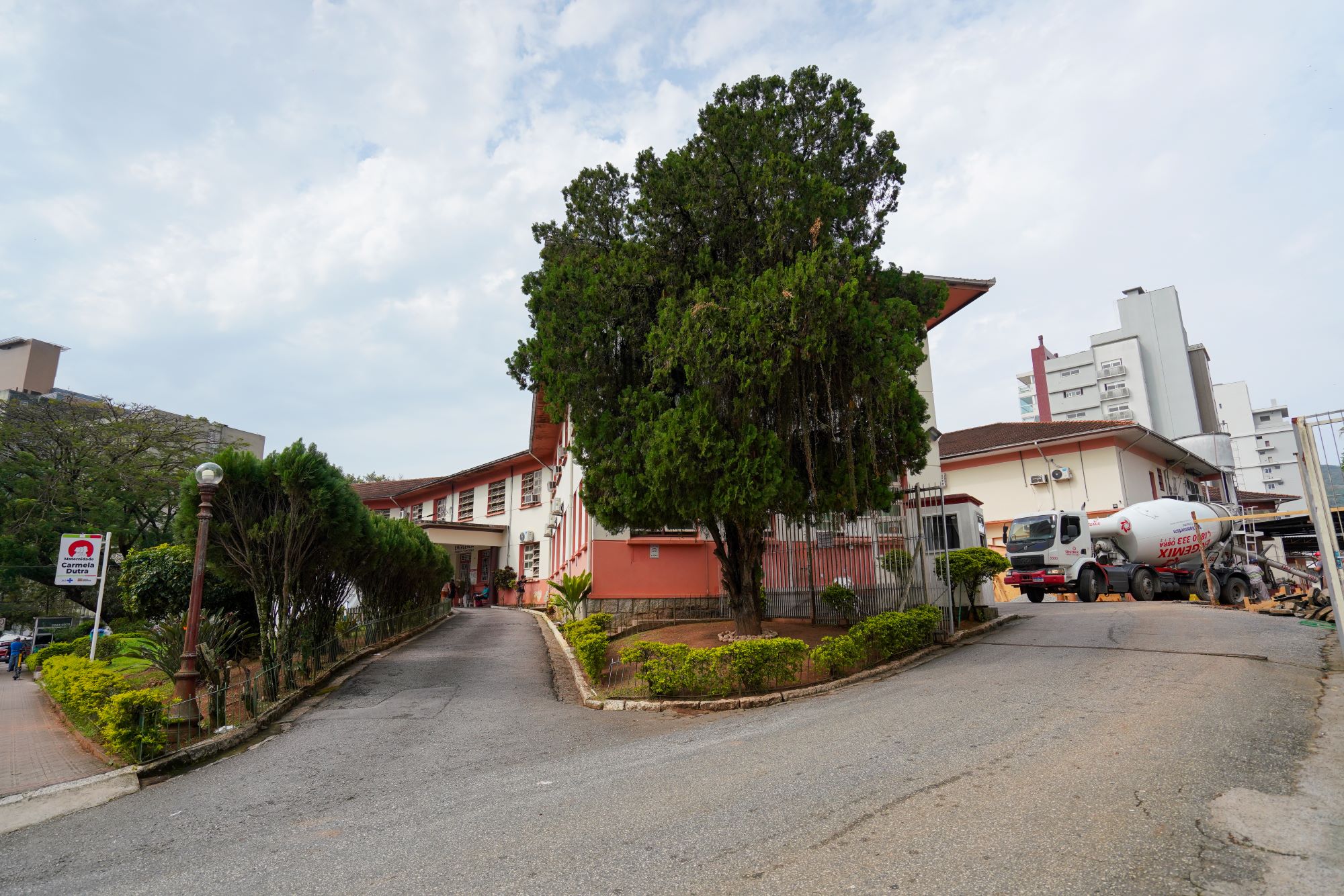 Vista externa da Unidade Carmela Dutra com rampa de acesso, jardim com árvores podadas, prédio de fachada vermelha ao fundo e caminhão betoneira estacionado à direita.