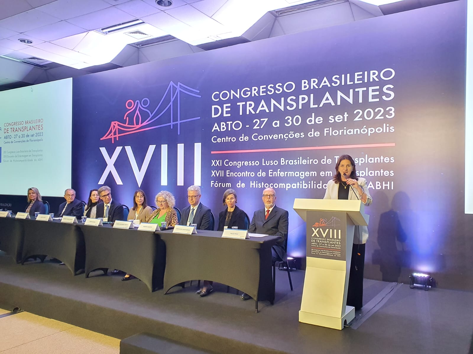 Fotografia de uma conferência com 11 pessoas sentadas em uma mesa no palco, e uma mulher discursando em um púlpito à direita. O fundo exibe o letreiro do XVIII Congresso Brasileiro de Transplantes – ABTO, realizado de 27 a 30 de setembro de 2023, no Centro de Convenções de Florianópolis. A iluminação destaca o púlpito e o painel principal com informações do evento.