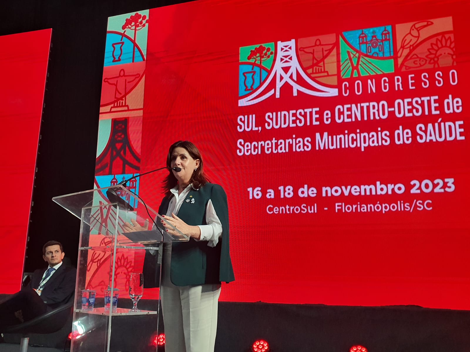 Mulher de pele clara e cabelos castanhos na altura dos ombros fala ao microfone em um púlpito de acrílico transparente, usando camisa branca e colete verde. Ao fundo, um grande telão vermelho exibe ilustrações coloridas de pontos turísticos e a frase 'Congresso Sul, Sudeste e Centro-Oeste de Secretarias Municipais de Saúde - 16 a 18 de novembro 2023 - CentroSul - Florianópolis/SC'. À esquerda, um homem de terno escuro e gravata está sentado, observando a palestra.