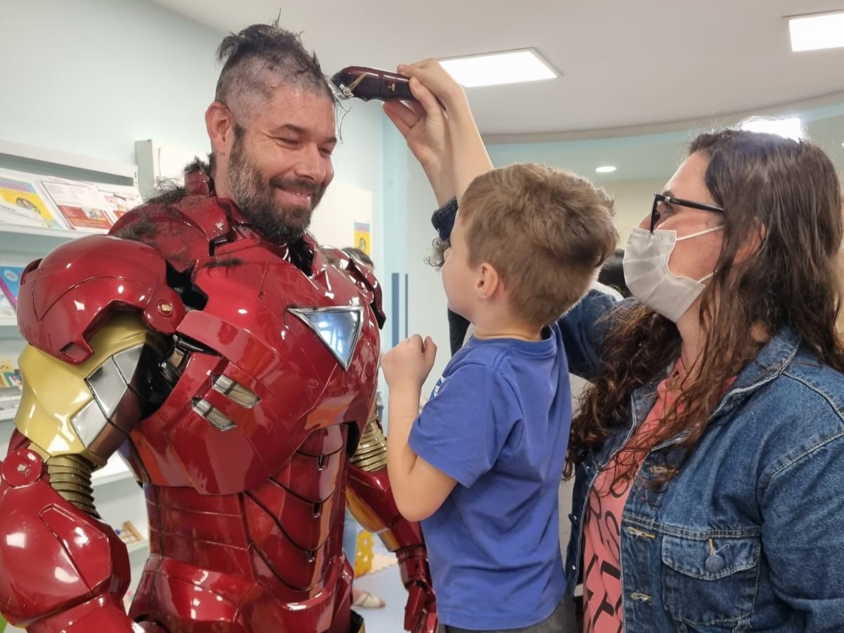 Homem vestido com fantasia do Homem de Ferro sorri enquanto um menino, no colo de uma mulher de óculos e máscara, usa uma máquina de cortar cabelo para raspar parte de sua cabeça. O ambiente é interno, com prateleiras e iluminação de teto.