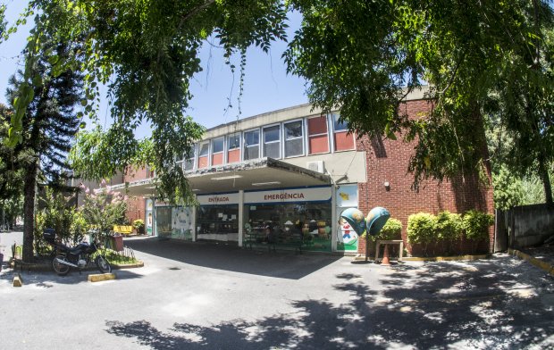 Fachada de um hospital com entrada de emergência sinalizada em português. O edifício tem paredes de tijolos vermelhos e janelas com vidros, parcialmente cobertas por uma marquise. Árvores ao redor projetam sombras no estacionamento, onde há uma motocicleta estacionada e esculturas coloridas de figuras infantis próximas à entrada. O ambiente é ensolarado, com céu azul visível entre as copas das árvores.