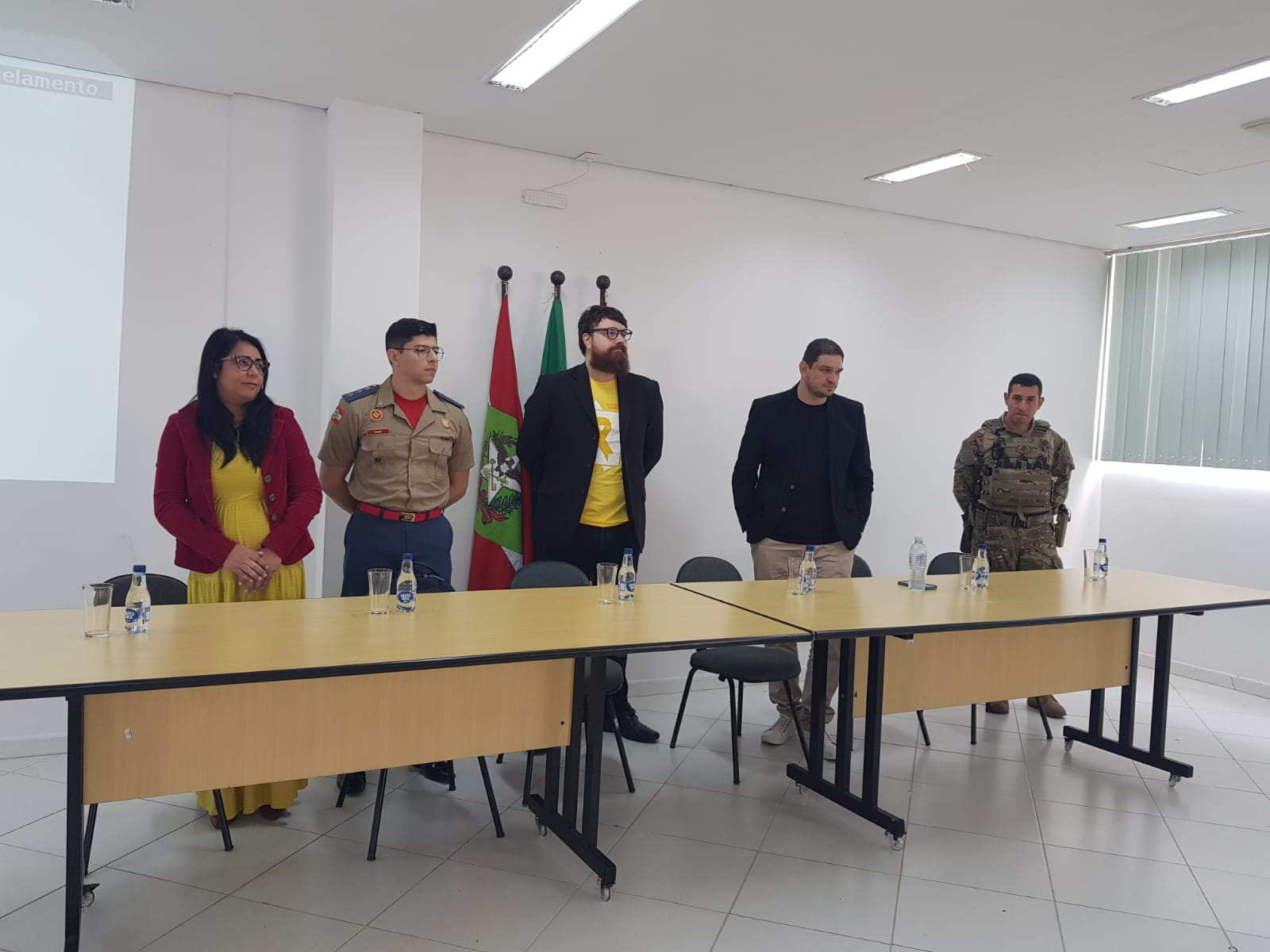 Cinco homens e uma mulher em pé atrás de uma mesa em ambiente de auditório. À esquerda, uma mulher de vestido amarelo e blazer vermelho está ao lado de um bombeiro militar. No centro, dois homens com camisetas amarelas e blazers escuros. À direita, um homem com blazer preto e outro com uniforme camuflado de forças de segurança. Ao fundo, bandeiras de Santa Catarina e do Brasil. Na mesa há garrafas de água e copos. Evento relacionado à prevenção ao suicídio.