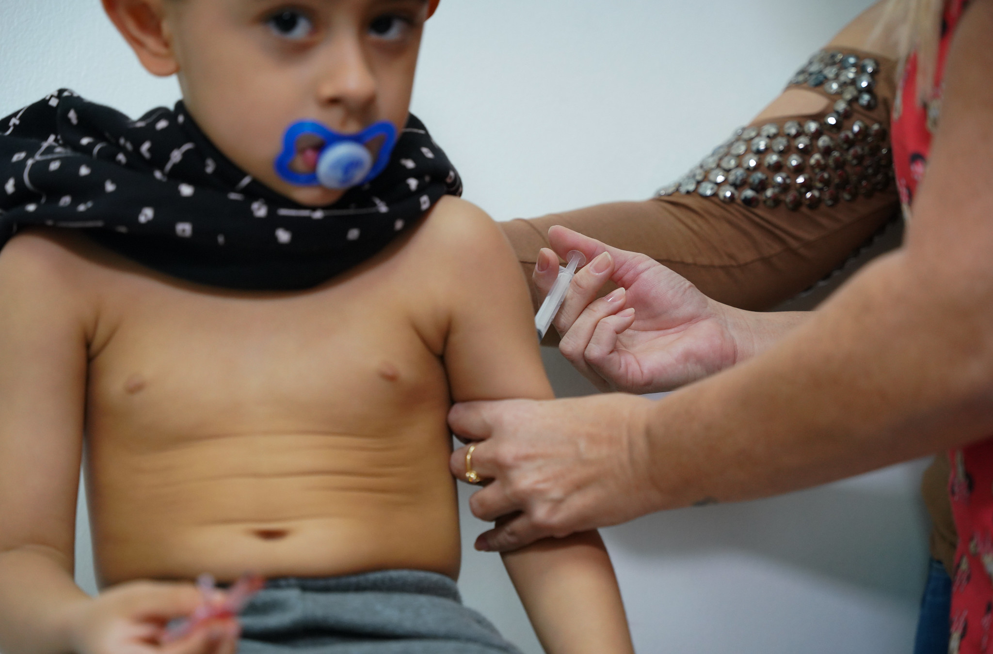 Menino pequeno, sem camisa e com chupeta na boca, recebe aplicação de vacina no braço por profissional de saúde, durante campanha de imunização.