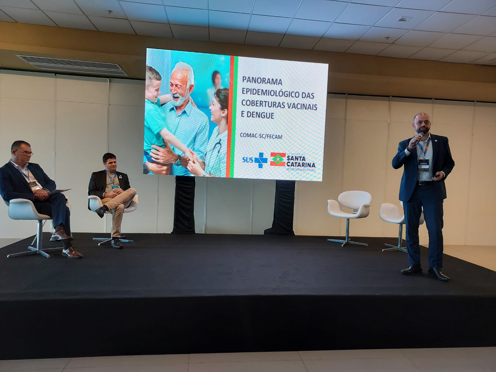 Apresentação em auditório com três homens, dois sentados à esquerda e um em pé à direita com microfone. Ao centro, uma tela exibe o tema “Panorama Epidemiológico das Coberturas Vacinais e Dengue”, com logotipos do SUS, Governo de Santa Catarina e Secretaria da Saúde. O palco é simples, com cadeiras brancas e ambiente corporativo.