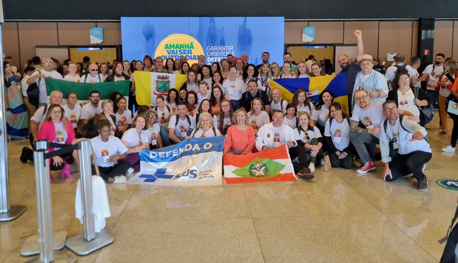 Grande grupo de pessoas posa sorrindo para foto em ambiente interno, durante a 17ª Conferência Nacional de Saúde. A maioria veste camisetas brancas com crachás e logotipos do evento. Algumas pessoas seguram bandeiras de diferentes estados e municípios, incluindo as de Santa Catarina, Rio Grande do Sul e do SUS (Sistema Único de Saúde), com os dizeres 'Defenda o SUS'. Ao fundo, há um painel azul com os dizeres 'Amanhã vai ser outro dia – Garantir direitos e defender o SUS'. O clima é de entusiasmo e união.