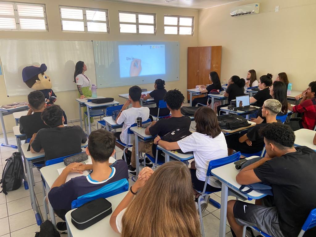 Sala de aula com cerca de vinte estudantes sentados em carteiras, atentos a uma apresentação feita por uma mulher em frente ao quadro branco. À esquerda da imagem, um mascote com uniforme e boné do SAMU está presente. A apresentação é projetada no quadro e o ambiente é iluminado por luz natural, com janelas superiores abertas. A ação faz parte do programa EducaSAMU, promovendo educação em saúde e primeiros socorros.