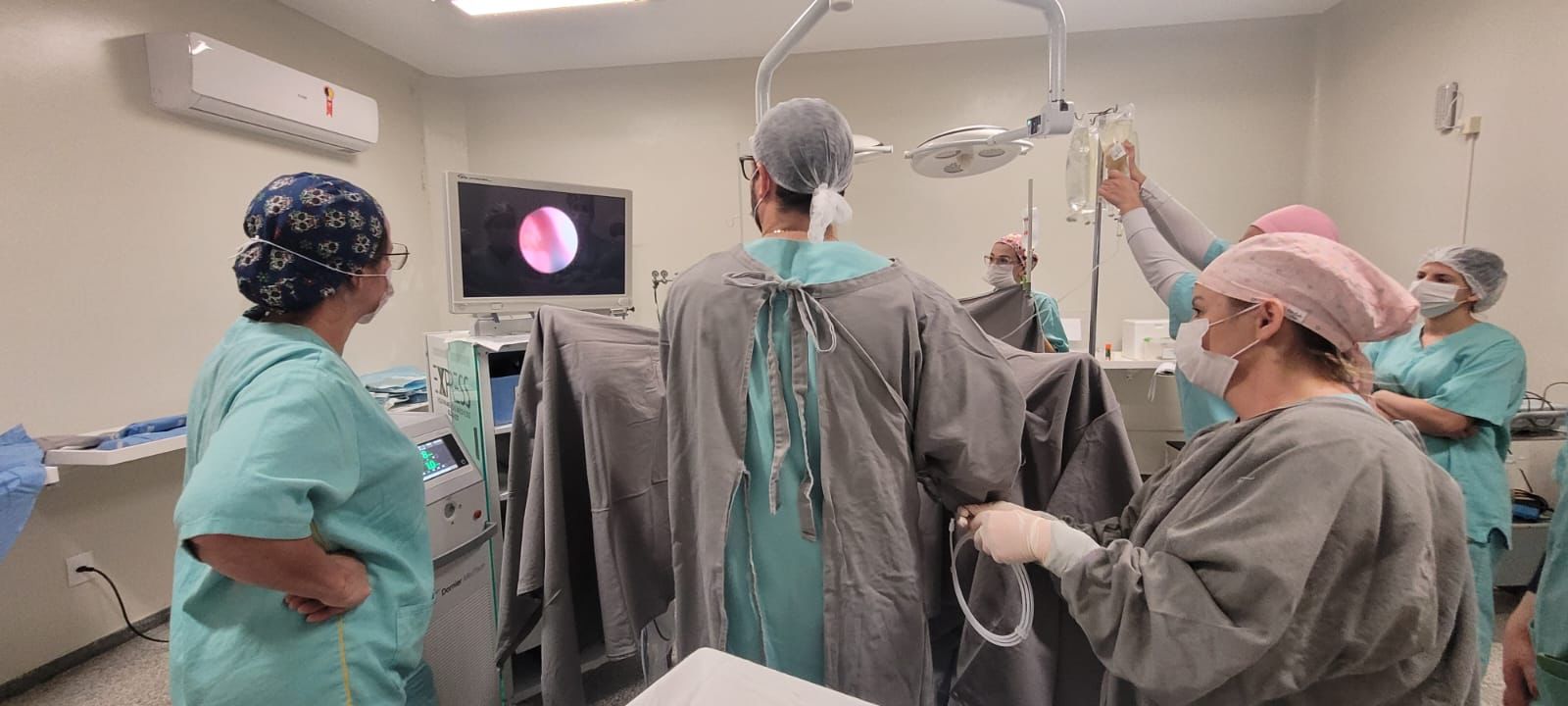 Equipe médica realiza procedimento por vídeo em centro cirúrgico. Profissionais observam a imagem endoscópica em um monitor, que mostra o interior do corpo do paciente. Alguns seguram bolsas de soro e outros manuseiam equipamentos. Todos usam roupas cirúrgicas, toucas e máscaras, em ambiente hospitalar estéril e iluminado.