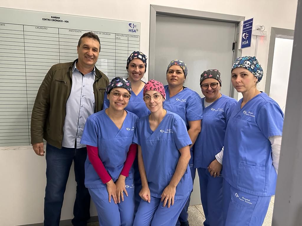 Seis profissionais da saúde, todas mulheres vestindo jaleco cirúrgico azul com toucas estampadas, posam ao lado de um homem com jaqueta marrom claro. Ao fundo, quadro Kanban do centro cirúrgico e porta cinza com a logo do Hospital Santo Antônio (HSA).