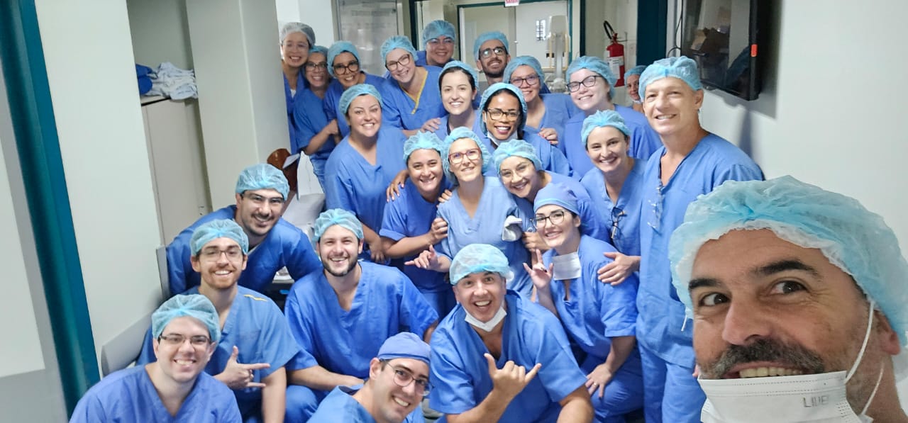 A imagem mostra um grupo numeroso de profissionais de saúde reunidos em um corredor de hospital, todos vestidos com roupas cirúrgicas azuis, toucas descartáveis e, em alguns casos, máscaras. Eles estão sorrindo e posando para a foto, transmitindo um clima de união e entusiasmo. A foto aparenta ter sido tirada antes ou depois de um mutirão cirúrgico, com os integrantes demonstrando espírito de equipe e colaboração.