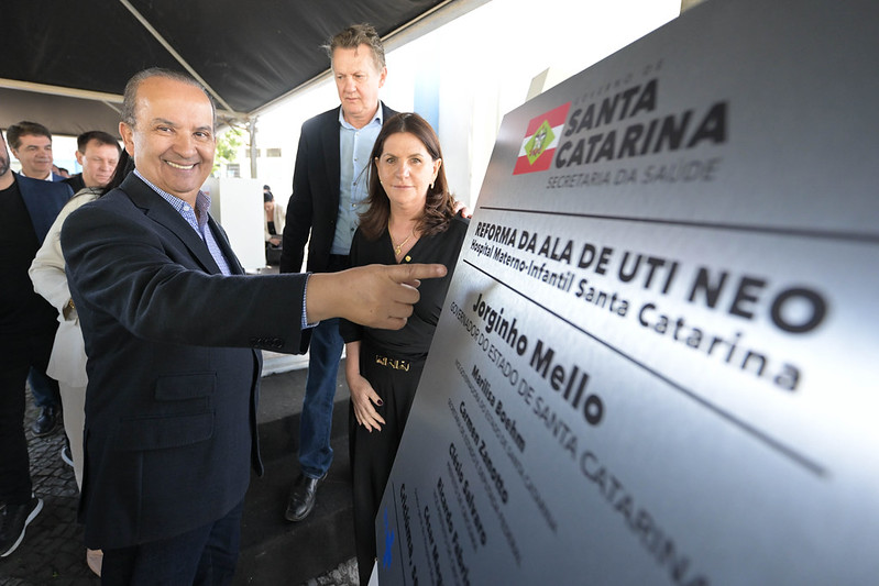 Governador Jorginho Mello sorri e aponta para placa comemorativa da reforma da ala de UTI Neonatal do Hospital Materno-Infantil Santa Catarina, acompanhado de autoridades sob tenda oficial.