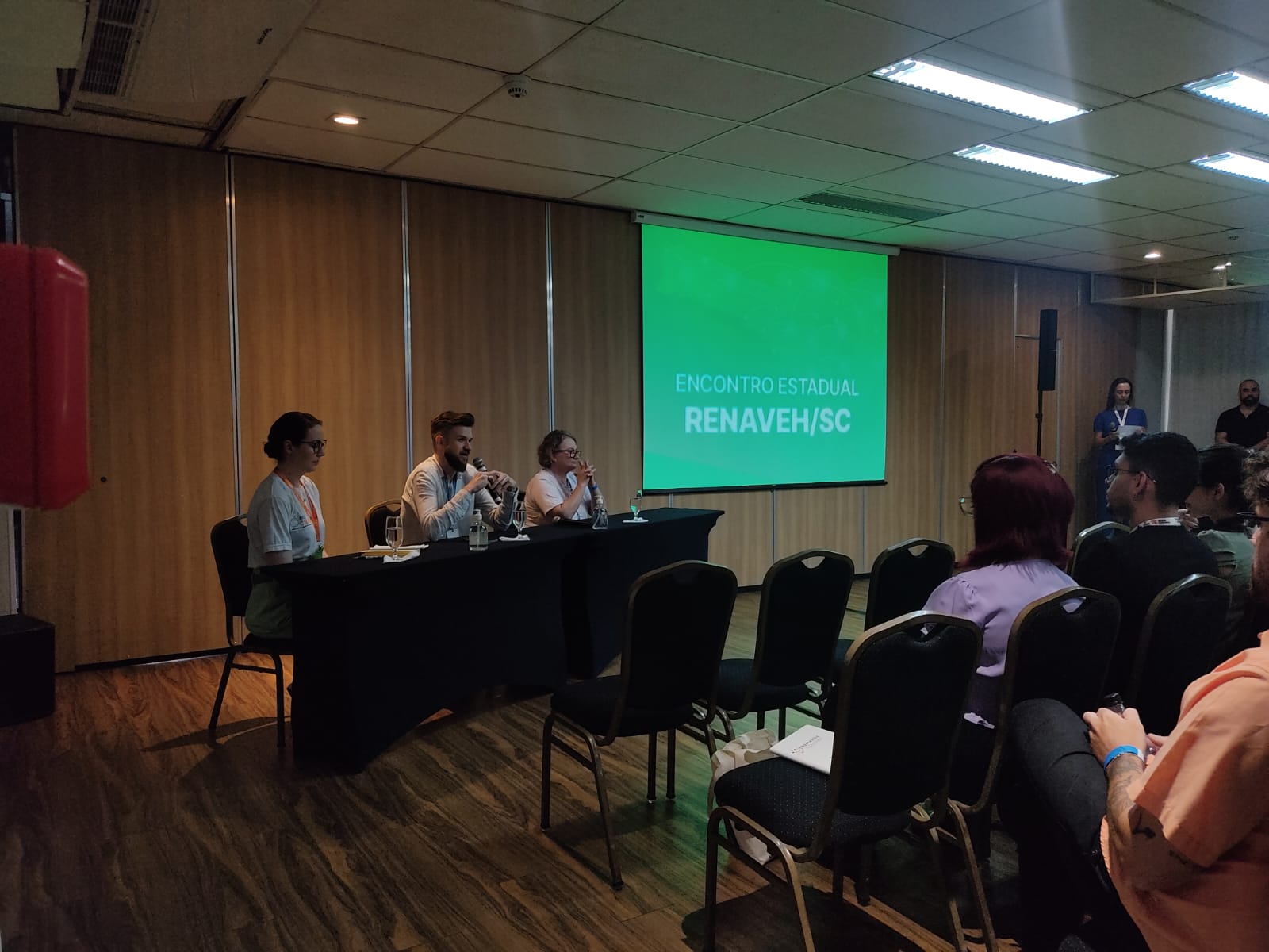 Três pessoas sentadas atrás de uma mesa preta participam de um painel de discussão, com microfones e copos de água à frente. Ao fundo, um telão verde exibe o texto “ENCONTRO ESTADUAL RENAVEH/SC”. Algumas pessoas assistem na plateia, em um auditório com piso de madeira e divisórias de madeira clara.
