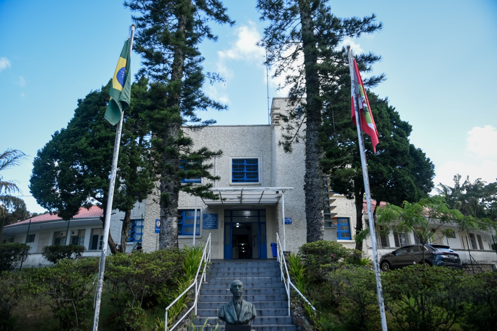 Fachada de um edifício público de dois andares, com janelas azuis e paredes de cor clara com textura áspera. Na entrada, há uma escadaria central ladeada por corrimãos brancos, vegetação ao redor e uma estátua de busto em homenagem a uma figura masculina. À frente da entrada principal, duas bandeiras estão hasteadas: a do Brasil e a de Santa Catarina. Árvores altas e vegetação completam o cenário externo.