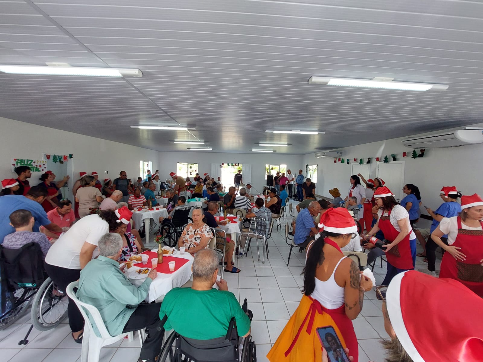 A imagem mostra uma confraternização de Natal no interior do Centro de Convivência do Hospital Santa Teresa.  O salão está decorado com enfeites natalinos nas paredes e mesas cobertas com toalhas brancas e vermelhas. Muitas pessoas, incluindo pacientes, alguns em cadeiras de rodas, e funcionários, estão reunidas.  Diversos participantes usam gorros vermelhos de Papai Noel, e alguns funcionários vestem aventais vermelhos, servindo alimentos e bebidas. O clima é festivo e acolhedor, com pessoas conversando, sorrindo e compartilhando a refeição. Ao fundo, há um cartaz na parede escrito “Feliz Natal”.