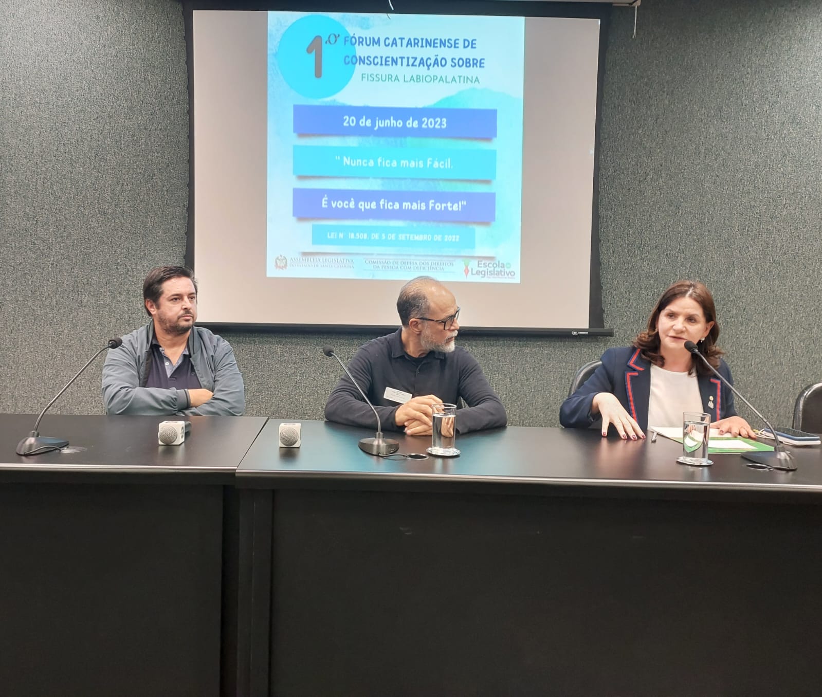 Três pessoas sentadas à mesa participam de painel durante o 1º Fórum Catarinense de Conscientização sobre Fissura Labiopalatina. Ao fundo, um telão projeta a data do evento (20 de junho de 2023) e a frase 'Nunca fica mais fácil. É você que fica mais forte!'. Uma das participantes, uma mulher à direita, fala ao microfone enquanto os dois homens ao lado observam.