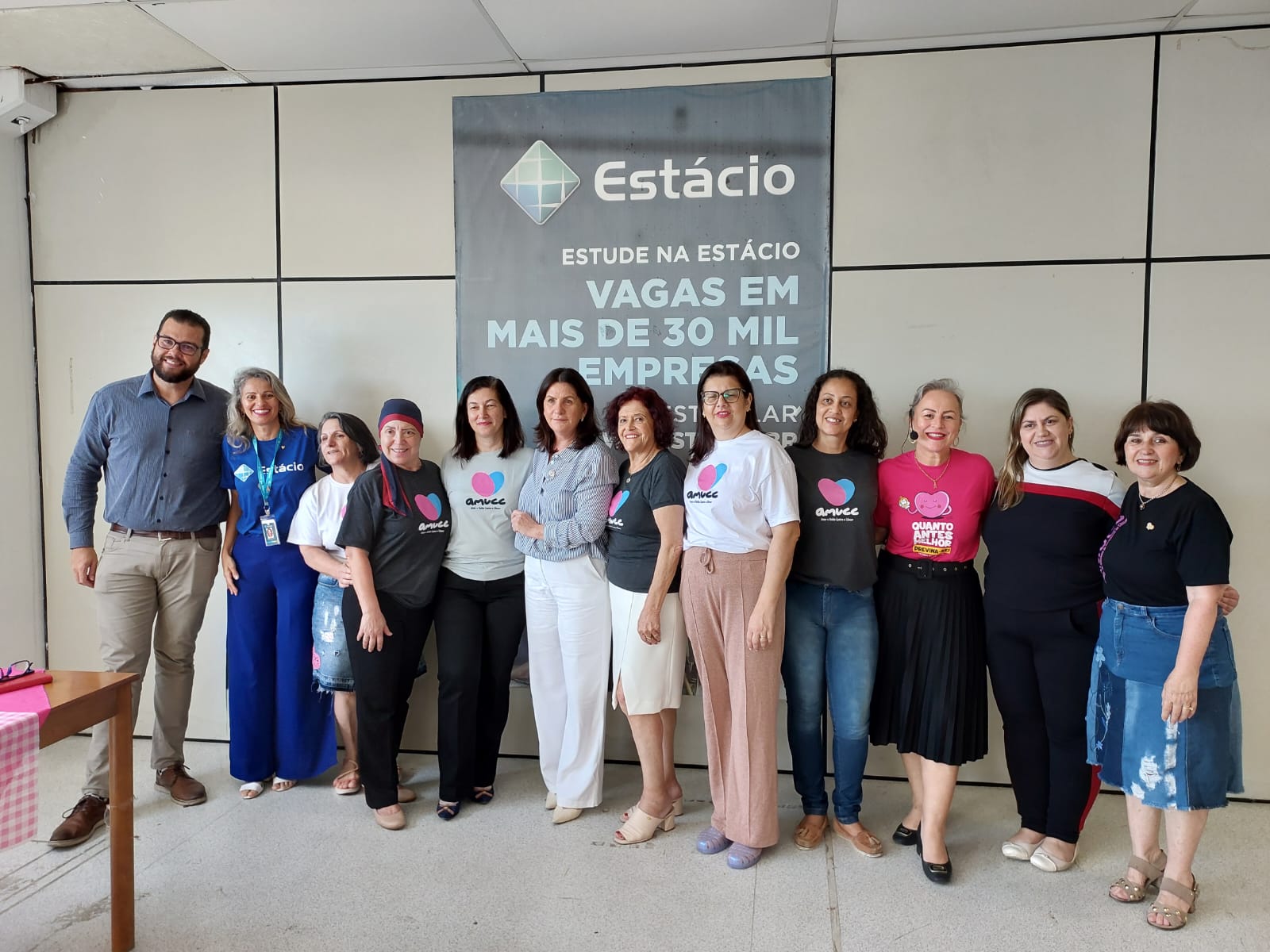 A imagem mostra um grupo de 12 pessoas posando para uma foto em frente a um painel da Estácio, que traz o texto “Estude na Estácio — Vagas em mais de 30 mil empresas”. Elas estão lado a lado, sorrindo, com alguns abraçando ou apoiando a mão nos ombros umas das outras, transmitindo clima de união e parceria.  As roupas variam, mas várias pessoas vestem camisetas da AMUCC (Associação Brasileira de Portadores de Câncer), com o logotipo de coração colorido. Também há uma participante usando camiseta rosa com a frase “Quanto mais cedo melhor”. À esquerda, um homem de camisa social azul e calça bege, e ao lado dele, uma mulher com uniforme azul da Estácio. O cenário é um espaço interno com paredes claras e piso claro, iluminado por luz natural.