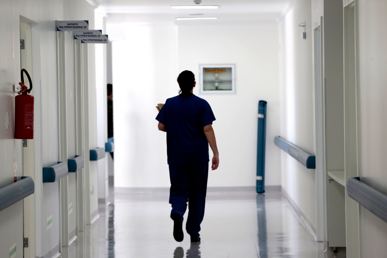 Pessoa vestida com uniforme azul de enfermagem caminha sozinha por um corredor hospitalar branco e bem iluminado. Nas paredes laterais há várias portas identificadas como 'Suporte Multiprofissional' e um extintor de incêndio visível à esquerda.