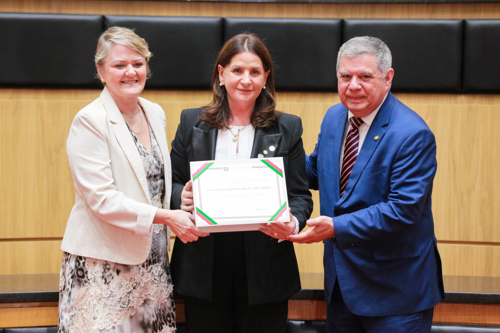 Três pessoas posam sorrindo em um auditório. A mulher ao centro segura um quadro com uma homenagem à Secretaria de Estado da Saúde de Santa Catarina, ladeada por um homem de terno azul e uma mulher com vestido estampado e blazer claro.