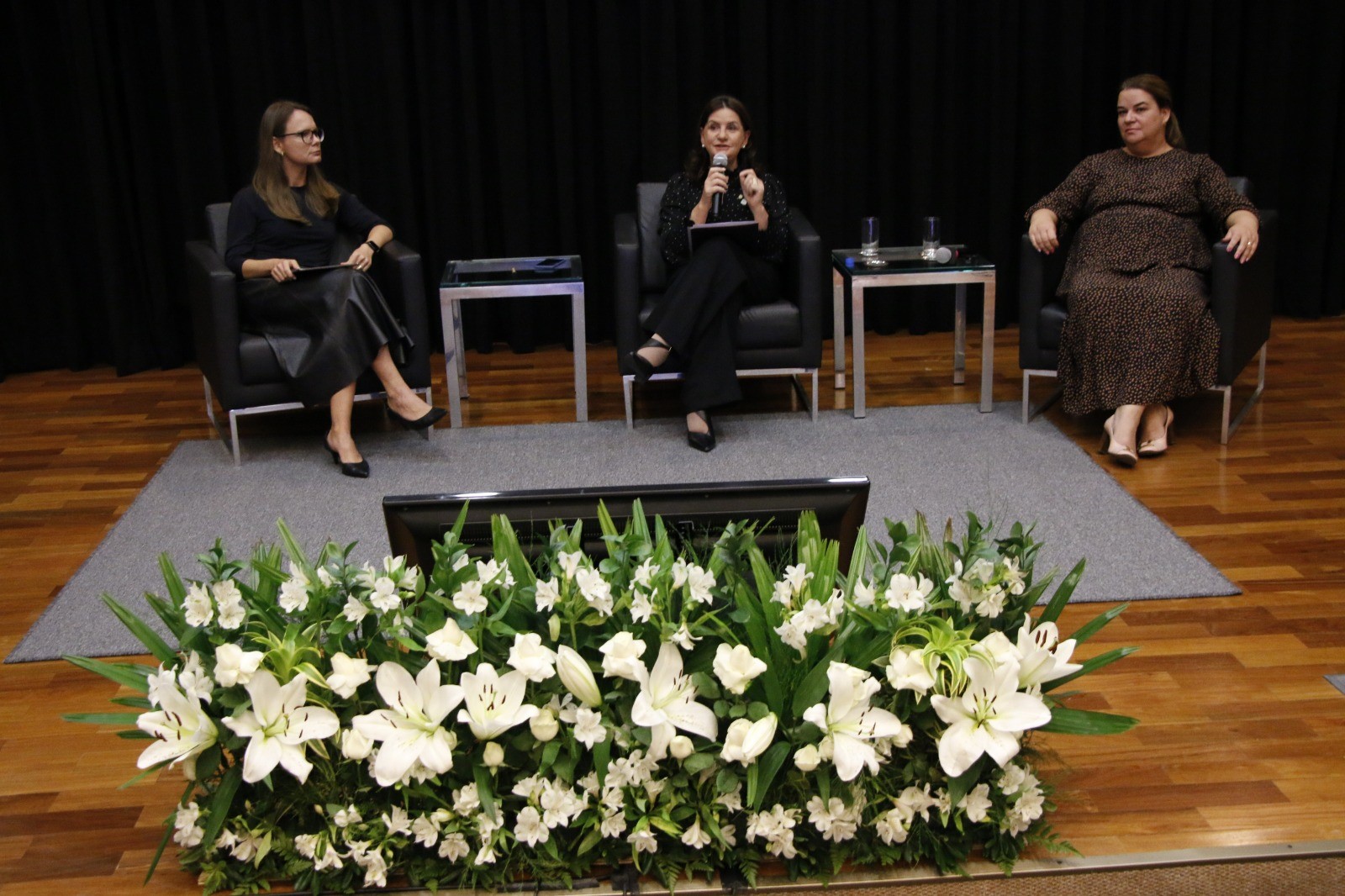 Três mulheres sentadas em poltronas sobre um palco durante um seminário. A mulher ao centro fala ao microfone. À frente do palco, há um arranjo floral com lírios brancos. O fundo é composto por uma cortina preta.