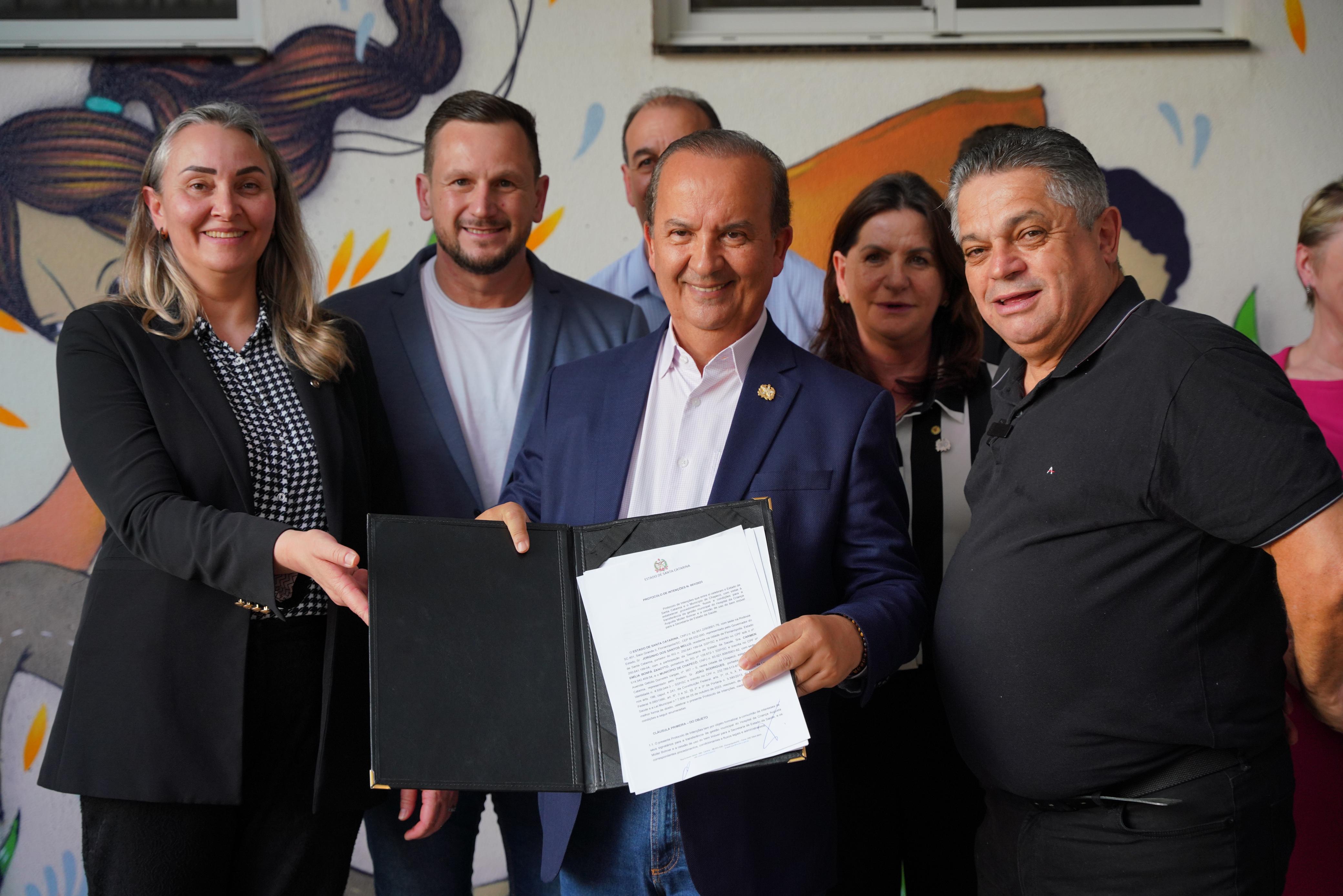 A imagem mostra um grupo de sete pessoas, sendo quatro em destaque na frente e três parcialmente visíveis ao fundo. O homem no centro segura uma pasta preta aberta, exibindo um documento oficial com timbre do Governo de Santa Catarina, enquanto sorri para a câmera.  As pessoas usam trajes formais ou semiformal, com exceção de um homem à direita vestindo camisa polo preta. O fundo é composto por um mural artístico colorido, com ilustrações de crianças e elementos lúdicos, sugerindo que o evento ocorre em um ambiente ligado à infância, possivelmente em um hospital ou instituição infantil.