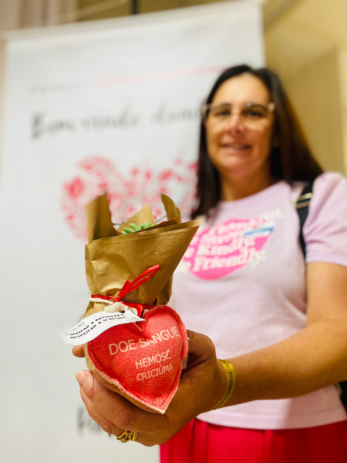 Mulher de camiseta rosa segura um pequeno vaso embrulhado em papel pardo, adornado com um coração de tecido vermelho com a inscrição “DOE SANGUE – HEMOSC CRICIÚMA”. Ao fundo, desfocado, há um banner branco com flores rosas e a frase “Bem-vindo doador”.