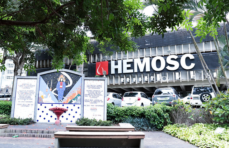 Fachada do HEMOSC (Centro de Hematologia e Hemoterapia de Santa Catarina), com letreiro grande em branco sobre fundo preto. À frente do prédio, há um mural artístico com mosaico colorido representando profissionais da saúde, além de trechos de texto nas laterais. Estacionamento com veículos e área com árvores e jardins compõem o entorno.