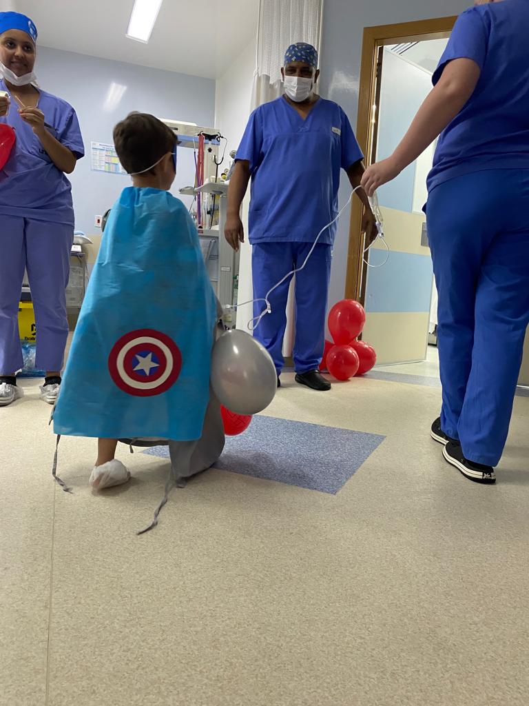 Criança vestida com capa azul do Capitão América caminha por um corredor hospitalar segurando balões vermelhos e prateados, acompanhada por profissionais de saúde em roupas cirúrgicas azuis. O ambiente é alegre e acolhedor, com clima de brincadeira antes ou depois de um procedimento médico.