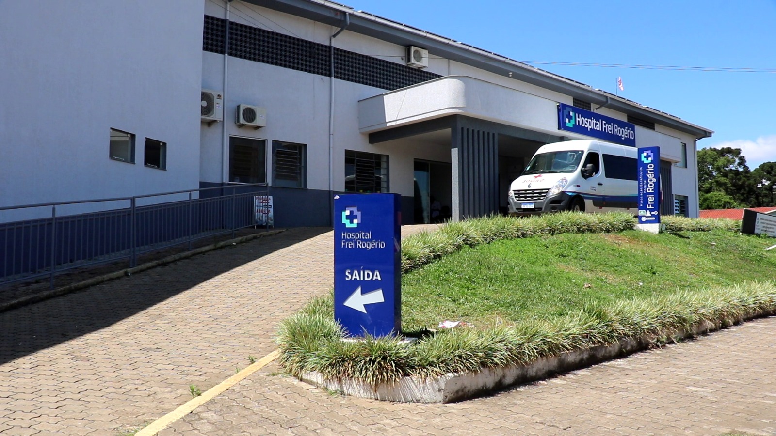 A imagem mostra a fachada do Hospital Frei Rogério, um prédio de cor clara com detalhes em cinza e telhado de duas águas. Na parte frontal, há uma rampa de acesso com corrimão e uma área coberta na entrada principal. Em frente ao prédio, um totem azul exibe o nome do hospital e a indicação de “SAÍDA” com uma seta apontando para a esquerda. À direita, uma ambulância branca está estacionada próxima à entrada. O céu está azul e ensolarado, sugerindo um dia claro, e há áreas gramadas e arborizadas ao redor do hospital.