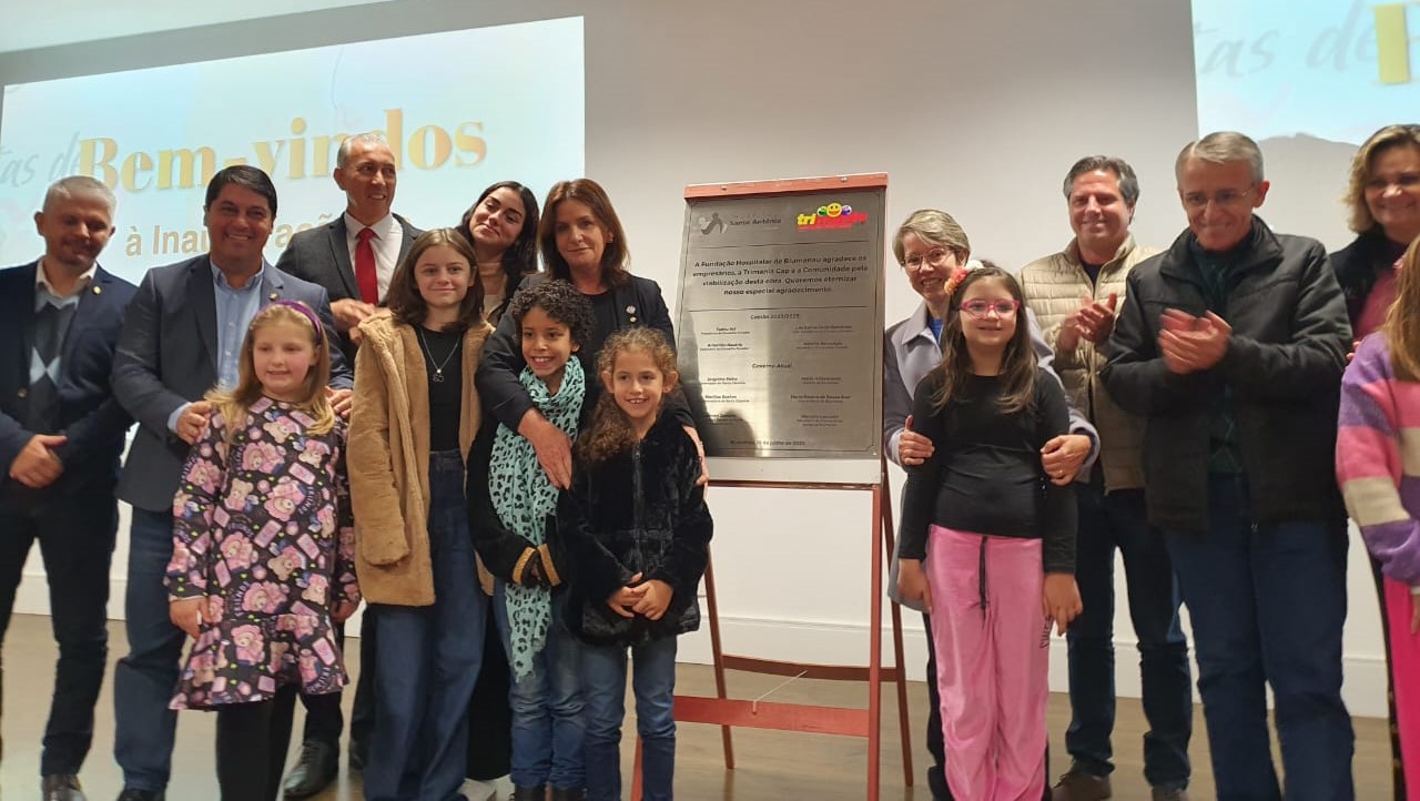 Grupo de autoridades e crianças reunidos ao redor de uma placa recém-inaugurada, com expressão de alegria e celebração. A cerimônia ocorre em ambiente interno, com um projetor ao fundo exibindo a palavra 'Bem-vindos'. Algumas pessoas aplaudem, enquanto outras posam para a foto em frente à placa que está sobre um cavalete.