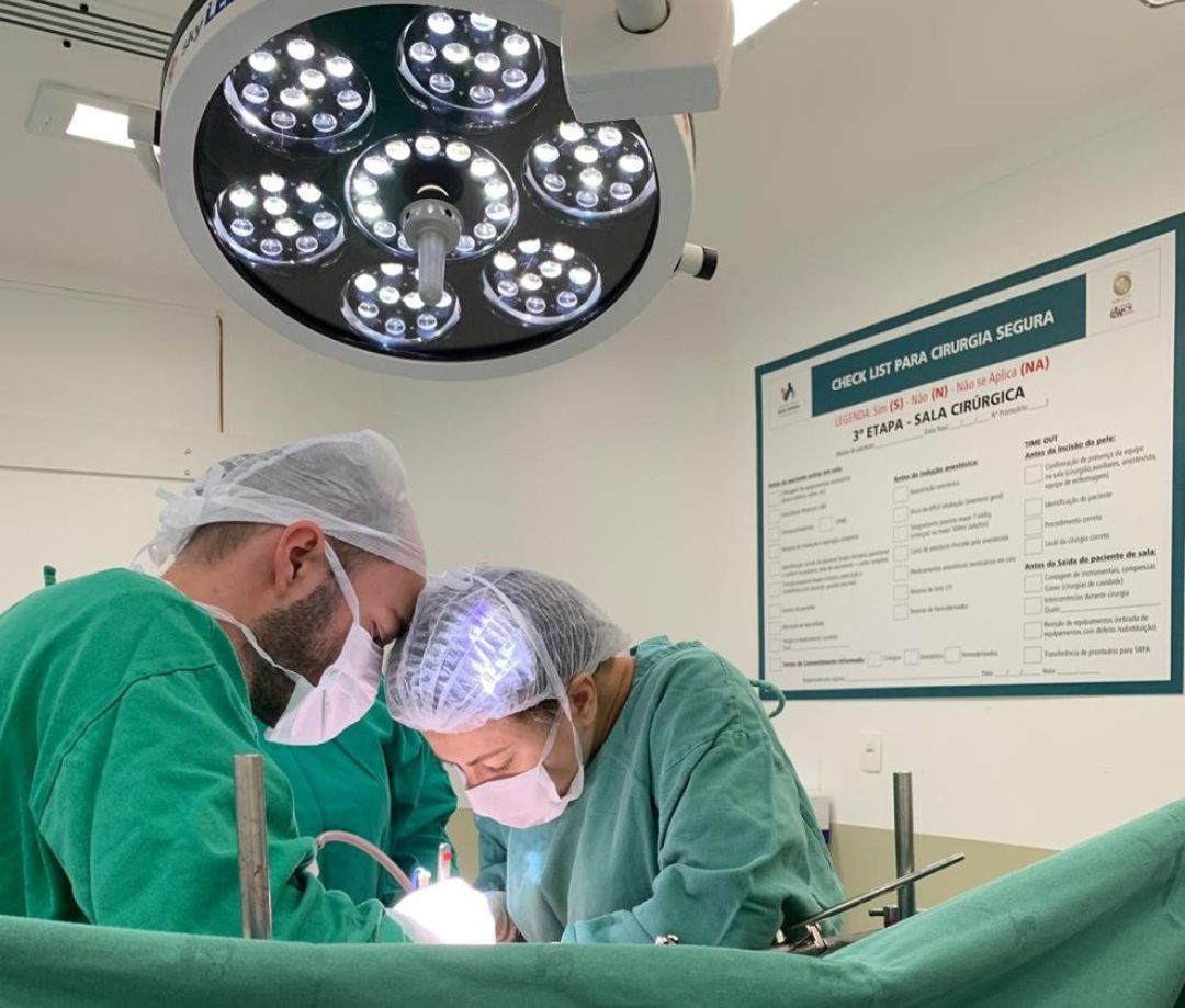 A imagem mostra uma cena dentro de uma sala cirúrgica.  Dois profissionais de saúde, vestidos com aventais, toucas e máscaras cirúrgicas, estão concentrados realizando um procedimento.  Acima deles há uma luminária cirúrgica com várias lâmpadas de LED acesas, iluminando intensamente o campo operatório.  Ao fundo, preso à parede, há um quadro de “Check List para Cirurgia Segura”, com campos para preenchimento e etapas do procedimento, reforçando protocolos de segurança.  A equipe aparenta estar em total foco e seguindo as normas de assepsia hospitalar. 