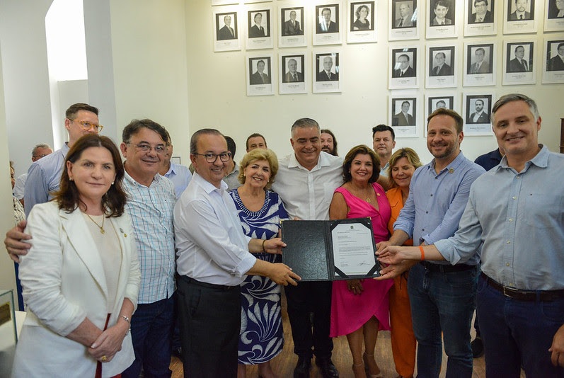 Grupo de autoridades e profissionais da saúde reunidos sorrindo para foto durante cerimônia de entrega de documento oficial no Hospital São José, em ambiente com parede decorada por retratos emoldurados.