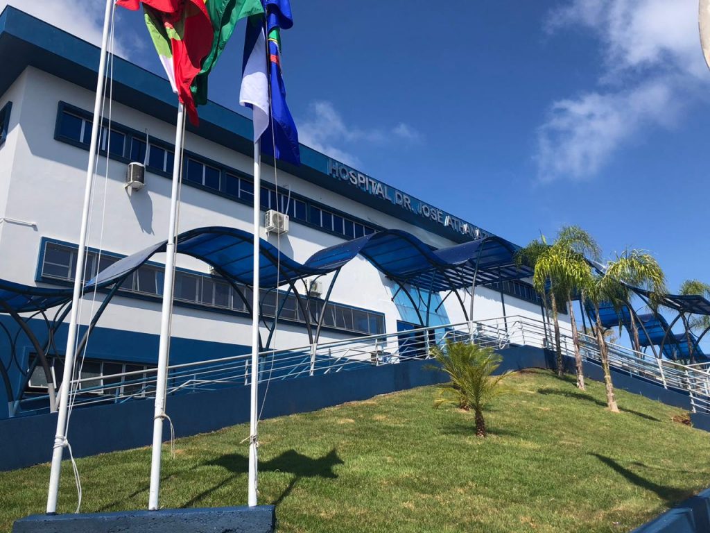 Fachada do Hospital Dr. José Athanázio em Campos Novos, com cobertura ondulada azul sobre rampa de acesso, bandeiras hasteadas e céu azul ao fundo.