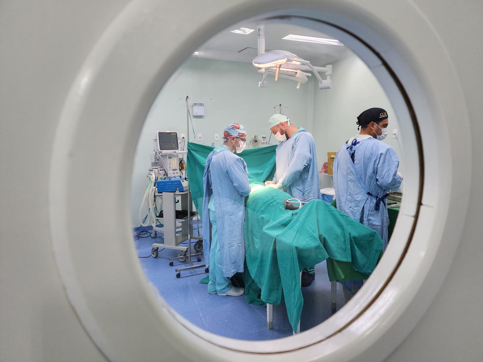 Cena observada por uma janela circular de porta, mostrando uma equipe de três profissionais de saúde vestidos com roupas cirúrgicas, gorros e máscaras, realizando um procedimento em um paciente coberto por campos cirúrgicos verdes, dentro de uma sala de cirurgia equipada com iluminação especial e aparelhos médicos.