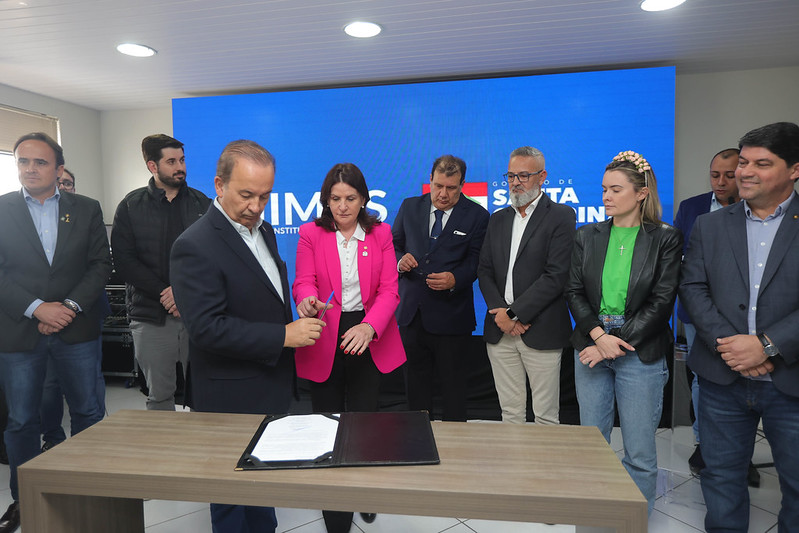 Cerimônia de assinatura com autoridades em frente a um painel digital azul com o logotipo do Governo de Santa Catarina e IMAS. Um homem assina um documento sobre uma mesa, enquanto outras pessoas observam, incluindo uma mulher de blazer rosa ao centro e diversos representantes em trajes formais ao fundo.