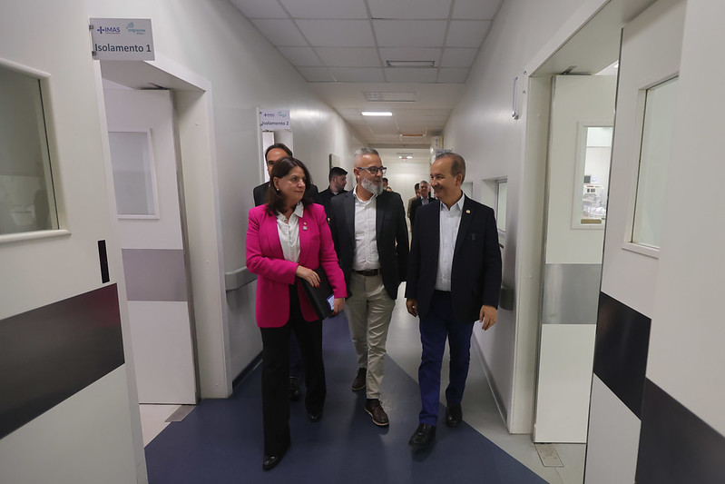 Autoridades caminham por um corredor hospitalar moderno durante uma visita institucional. À esquerda, uma mulher com blazer rosa conversa com dois homens de terno. As portas nas laterais estão identificadas como ‘Isolamento 1’ e ‘Isolamento 2’, e o ambiente é limpo e bem iluminado.