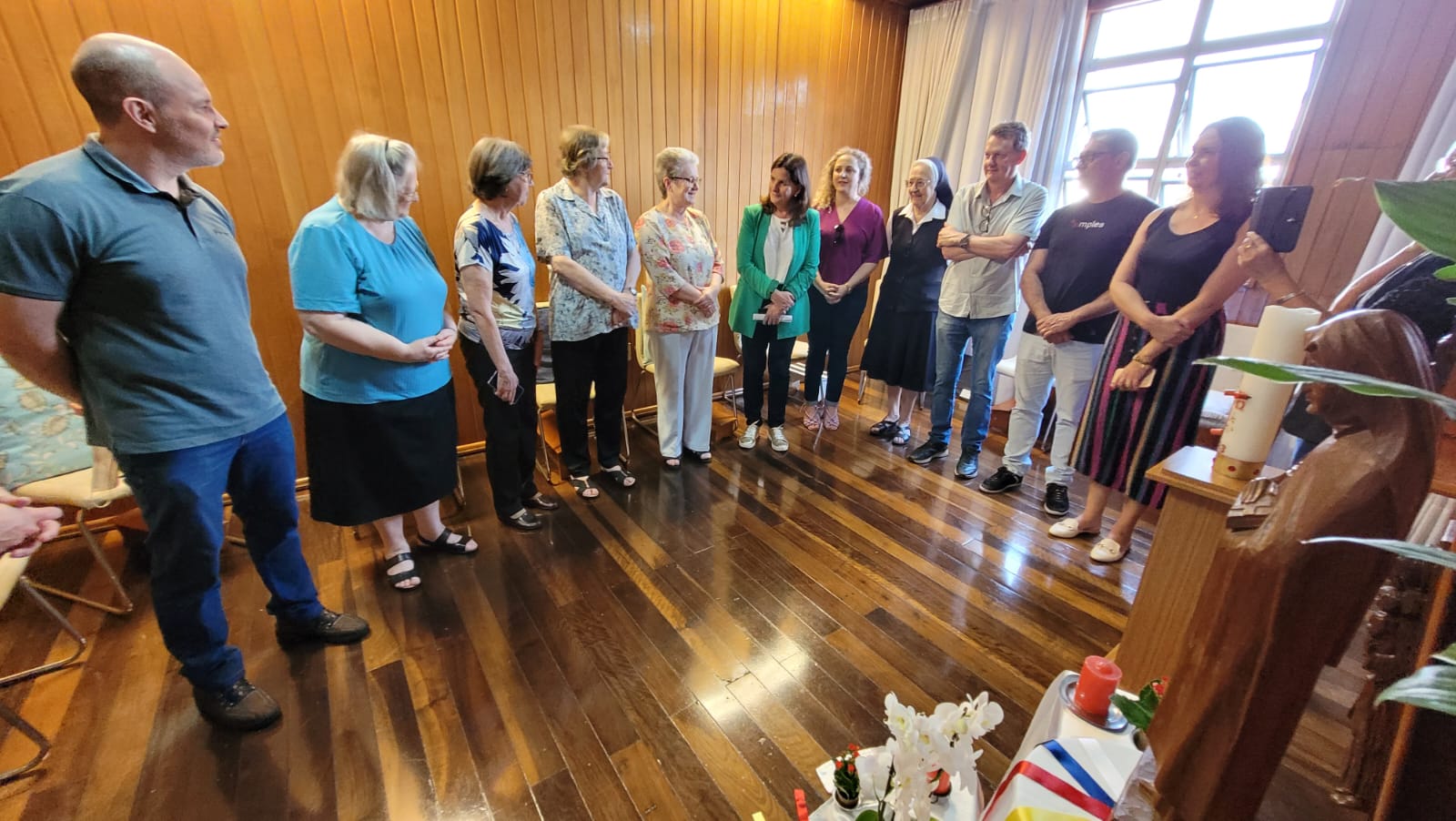 Grupo de cerca de 14 pessoas em pé, formando um semicírculo em uma sala com piso e paredes de madeira. A maioria das pessoas está sorrindo e conversando, incluindo religiosas e representantes da área da saúde. No canto inferior direito, há uma pequena mesa com velas, flores brancas e uma imagem religiosa, sugerindo que o local seja uma capela ou ambiente de espiritualidade dentro de um hospital.