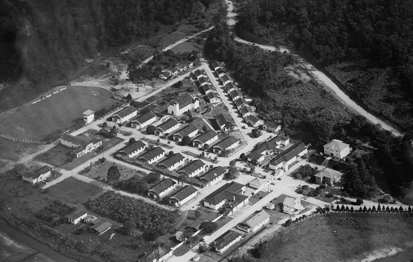 Fotografia aérea em preto e branco de um complexo hospitalar com vários prédios organizados em formato triangular, cercado por vegetação e estrada de terra. À esquerda, um campo de futebol pode ser visto. A imagem mostra a estrutura do hospital e sua integração com o ambiente natural ao redor.